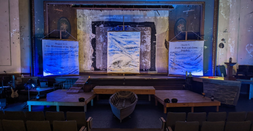 A theater stage with blue lighting, featuring large painted backdrops and a wooden boat in the foreground.