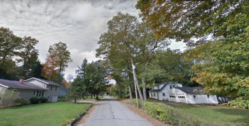 A quiet residential street lined with trees and houses, featuring a cloudy sky and autumn foliage.