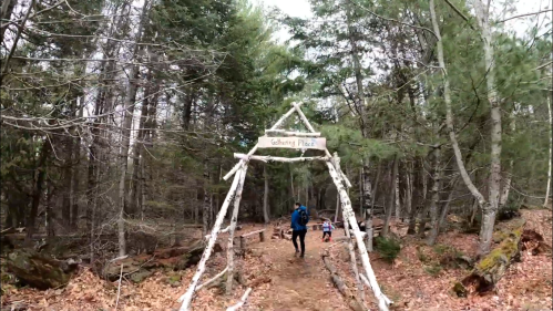 A person walks under a wooden archway labeled "Gathering Place" in a forested area with fallen leaves.