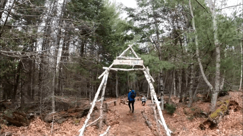 A person walks under a wooden archway labeled "Gathering Place" in a forested area with fallen leaves.