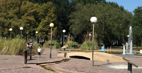 A park scene with children playing near a fountain, surrounded by greenery and lamp posts on a sunny day.