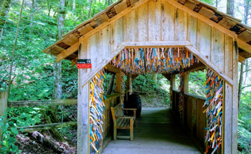 A wooden bridge entrance surrounded by trees, decorated with colorful ribbons and a bench inside.