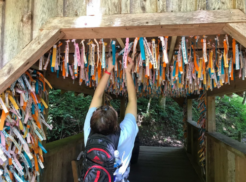 A person reaches up to touch colorful ribbons hanging from a wooden structure in a forested area.
