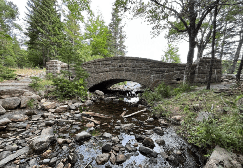 A stone arch bridge spans a shallow stream, surrounded by trees and rocky terrain.