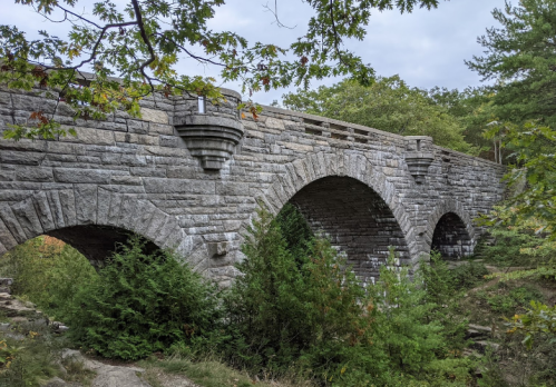 Stone bridge with arches, surrounded by greenery and trees, under a cloudy sky.