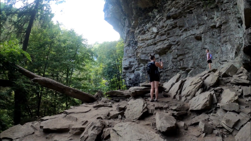 Two hikers explore a rocky trail beneath a large cliff, surrounded by lush green trees.