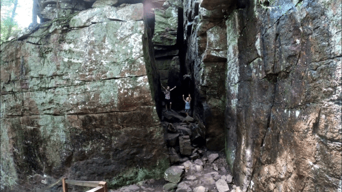 Two people stand in a rocky cave entrance surrounded by tall, moss-covered stone walls.