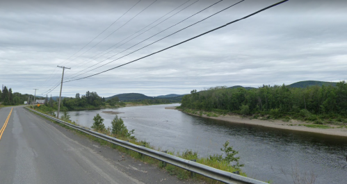 A winding river bordered by trees and hills, with a road and power lines alongside under a cloudy sky.