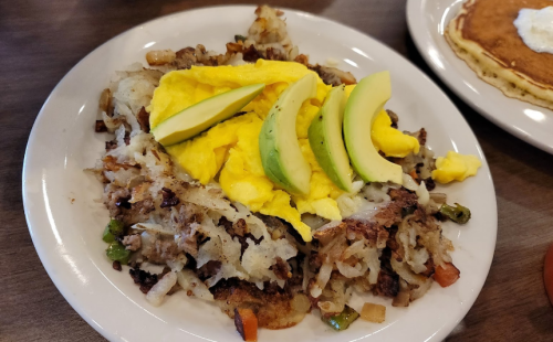 A plate of hash browns topped with scrambled eggs and avocado slices, served alongside pancakes.
