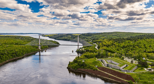 Aerial view of a cable-stayed bridge over a river, surrounded by lush greenery and a cloudy blue sky.