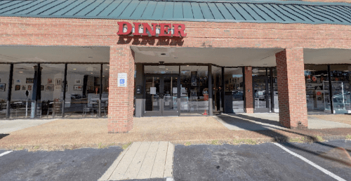 Exterior view of a diner with a red sign, large glass windows, and a brick facade, located in a shopping area.