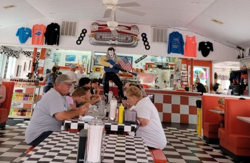 A retro diner with checkered floors, colorful decor, and patrons enjoying meals at a long table. Elvis statue in the background.