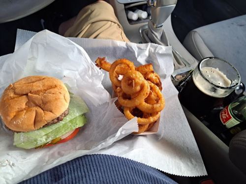 A burger with lettuce and tomato, curly fries, and a glass of dark soda on a car seat.