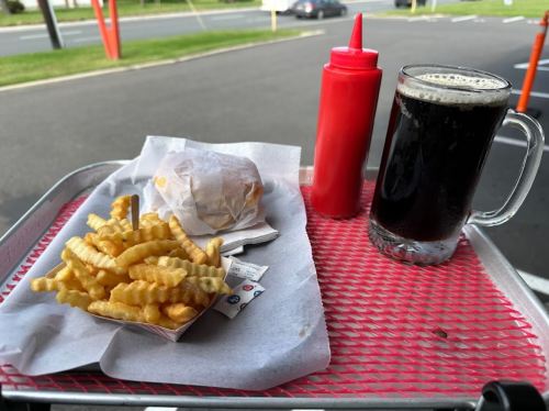 A tray with crinkle-cut fries, a burger, ketchup, and a mug of dark soda on a red picnic table.