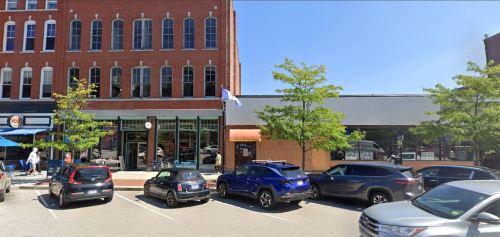 A sunny street scene featuring a brick building, parked cars, and trees lining the sidewalk.