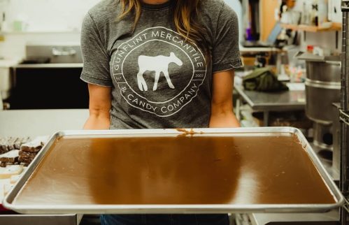 A smiling woman in a gray shirt holds a tray of chocolate in a candy shop kitchen.