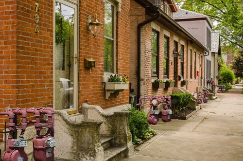 A row of brick houses with gas meters lined along the sidewalk, featuring planters and greenery.