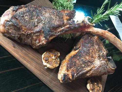 A wooden platter featuring two large, seasoned cuts of meat with roasted garlic and fresh herbs.