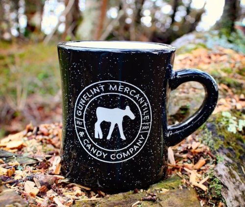 A black mug with a cow logo and text reading "Gunkel's Mercantile & Candy Company" on a log in a forest setting.