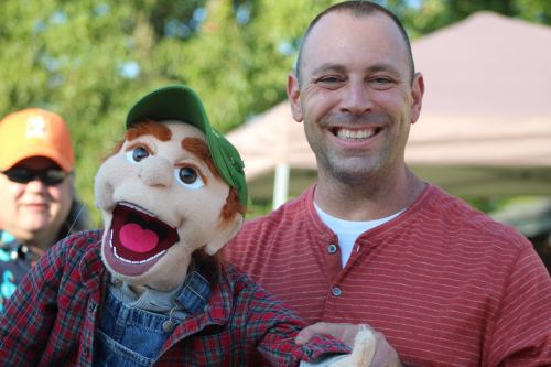 A smiling man in a red striped shirt holds a puppet with a green cap, both enjoying a sunny outdoor event.