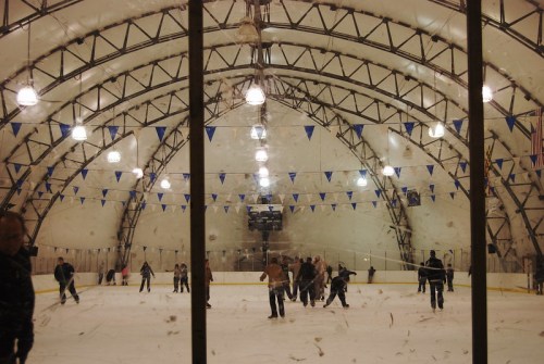 A busy indoor ice rink with skaters, bright lights, and blue pennants hanging from the ceiling.