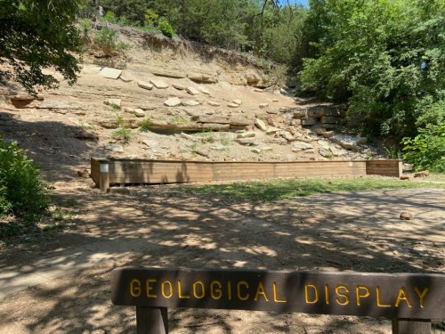 A wooden geological display in front of a rocky hillside, surrounded by trees and greenery.