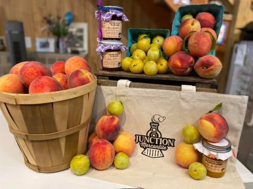A wooden basket filled with peaches, jars of jam, and a tote bag with "Junction Mercantile" logo, all on a table.