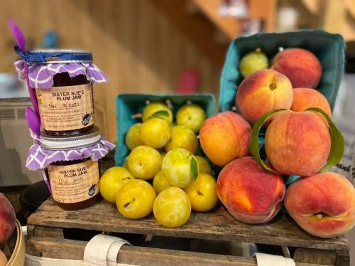 Jars of plum jam beside fresh yellow plums and ripe peaches on a wooden display.