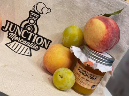A jar of peach jam with fresh peaches and plums, set against a Junction Mercantile tote bag.