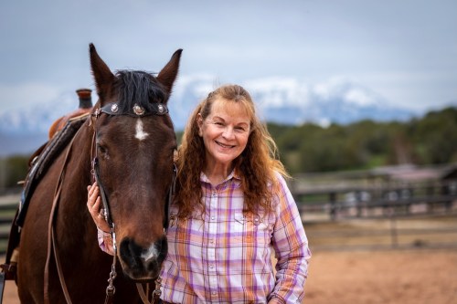 A smiling woman with long, curly hair stands beside a brown horse, with mountains in the background.