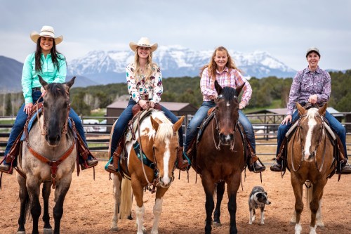 Four women on horseback, wearing cowboy hats, with mountains in the background and a dog nearby.