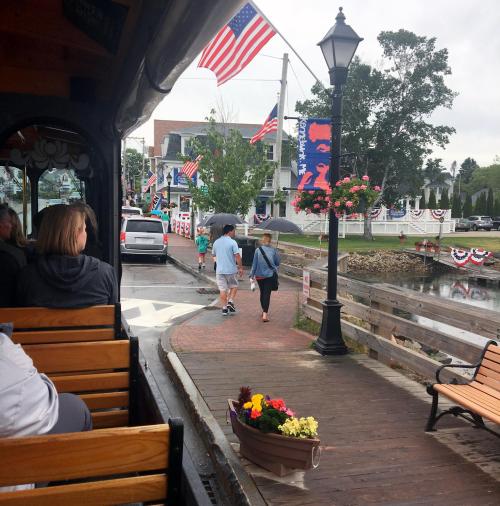 View from a trolley showing a decorated street with American flags, pedestrians, and a waterfront scene.