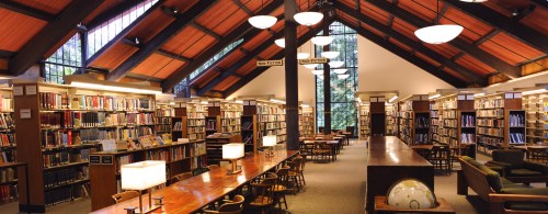 A spacious library with wooden beams, bookshelves, and tables, illuminated by warm lighting and large windows.