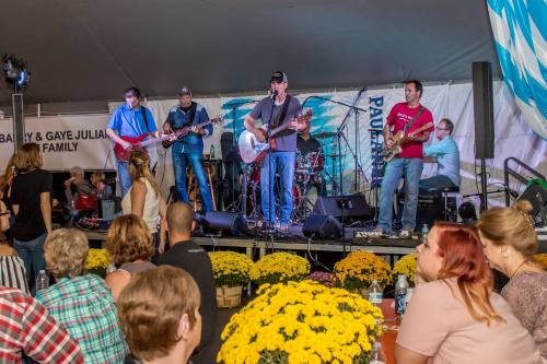 A live band performs on stage while an audience enjoys the show, surrounded by colorful flowers.