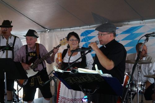 A band performs on stage, featuring musicians in traditional attire, with a woman in a dirndl and a man speaking into a microphone.