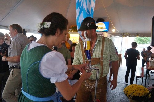 A woman in traditional Bavarian attire pours beer from a tap, surrounded by festival-goers under a tent.