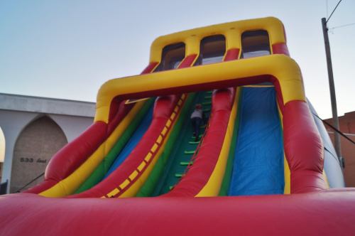 A colorful inflatable slide with three lanes, set against a clear sky. A child is climbing up the slide.