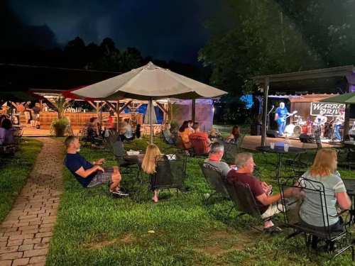 A lively outdoor music event at night, with people seated on lawn chairs enjoying a band performance under a tent.