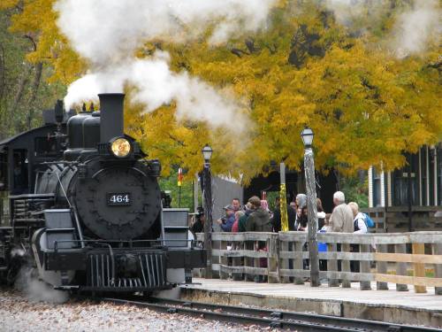 A steam train emits smoke near a crowd of people by a wooden fence, with vibrant yellow trees in the background.