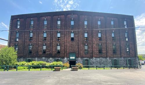 A large, brick building with multiple windows and a green entrance, surrounded by greenery and a clear blue sky.