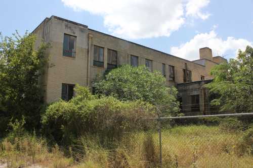 Abandoned building overgrown with vegetation, featuring broken windows and a chain-link fence in the foreground.