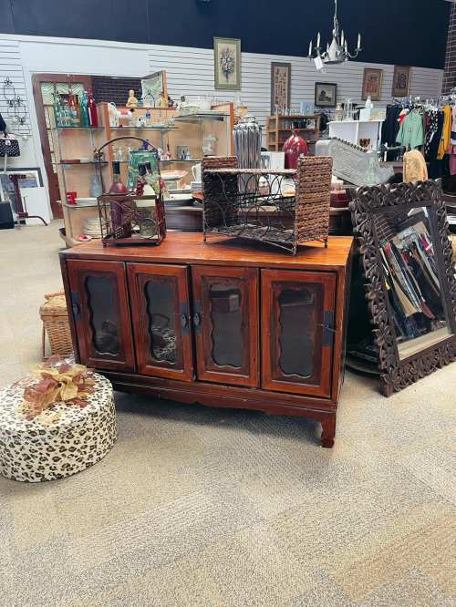 A wooden cabinet with glass doors sits in a vintage store, surrounded by various decor items and furniture.