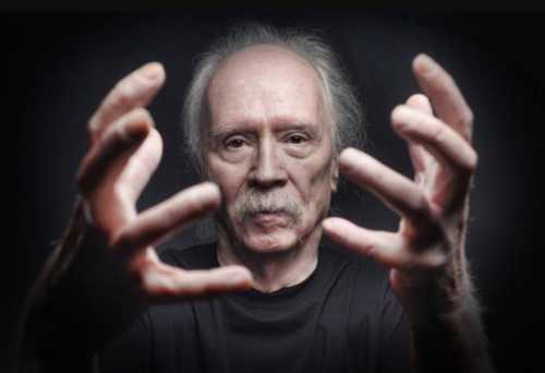 A man with gray hair and a mustache extends his hands forward, set against a dark background.