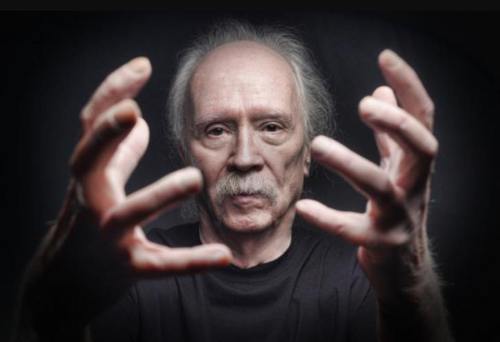 A man with gray hair and a mustache extends his hands forward, set against a dark background.