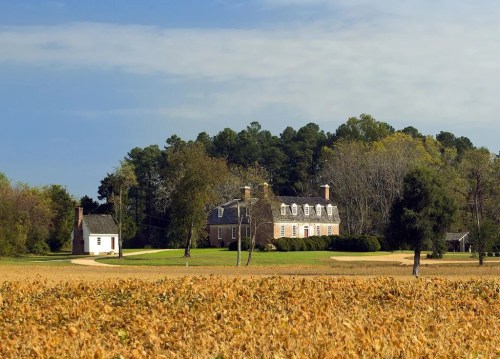 A large historic house with a smaller building nearby, set against a backdrop of trees and a golden field.
