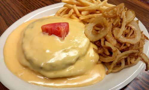 A plate featuring a creamy cheeseburger topped with a tomato slice, served with fries and crispy onion rings.
