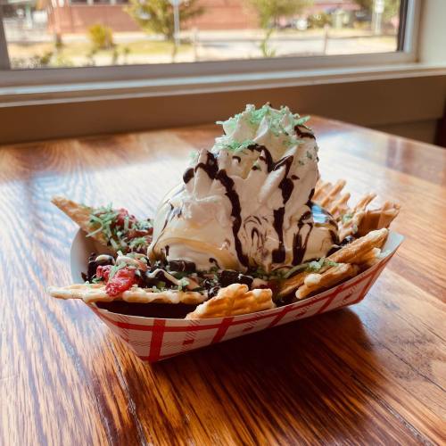 A dessert bowl with waffle fries topped with ice cream, chocolate sauce, and whipped cream, set on a wooden table.