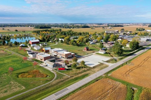 Aerial view of a rural town with fields, a pond, and various buildings, showcasing a peaceful landscape.