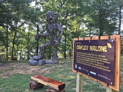A wooden statue of a forest creature stands beside an informational sign about Oakley Malone in a wooded area.