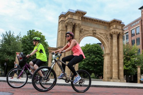 Two cyclists ride past a large stone archway surrounded by trees and buildings on a sunny day.